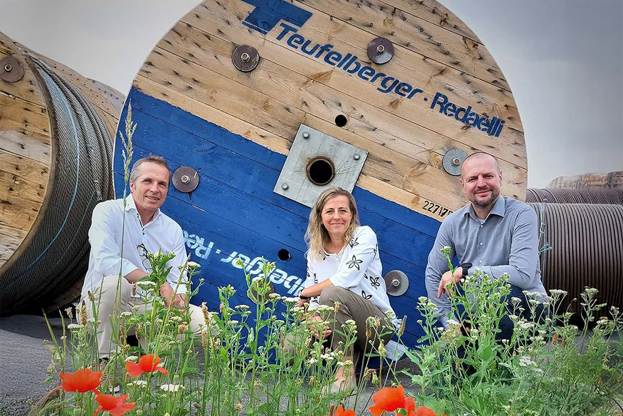 Three people sitting in front of a large wooden spool labeled “Teufelberger-Redaelli”. Flowers and greenery are visible in the foreground, with additional cable spools in the background.