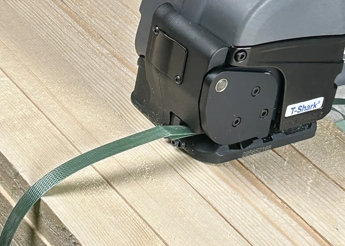 A close-up image of a T-Shark strapping tool securing a green strap around wooden planks. The tool is black and gray with the label "T-Shark" visible on its side.