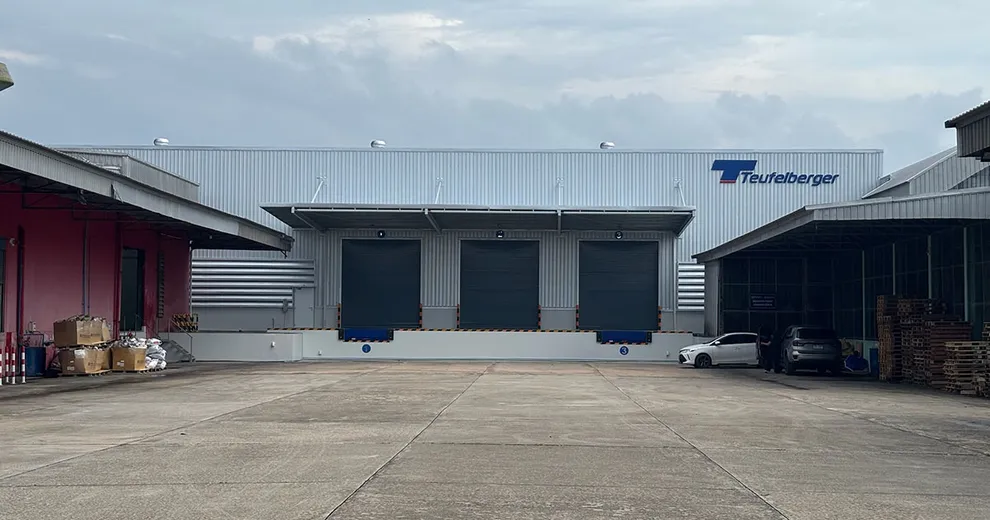 Industrial warehouse with three loading bays, marked "Teufelberger." A few parked vehicles and stacked pallets are visible. Overcast sky above.