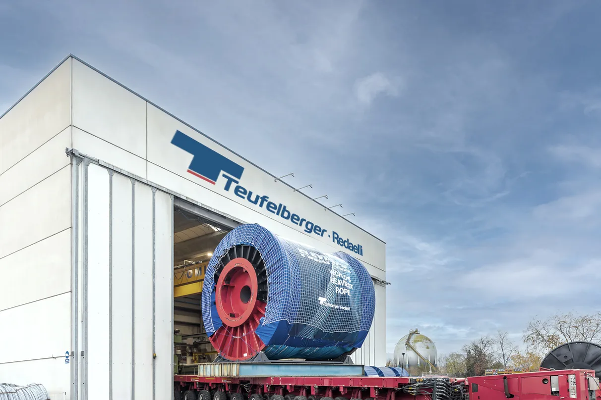A large industrial building labeled “Teufelberger - Redaelli” with a massive spool of cable wrapped in blue material, mounted on a red transport vehicle. Clear sky and trees in the background.