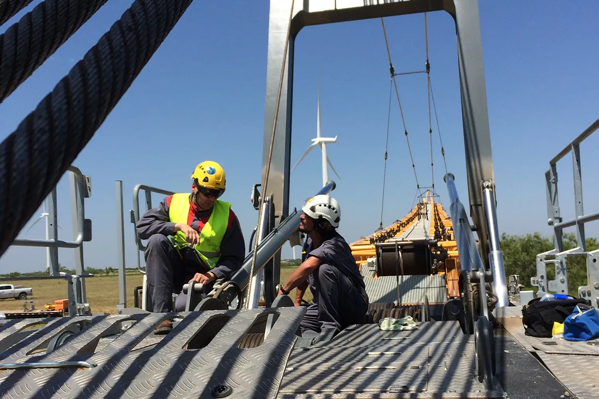Two workers wearing helmets and reflective vests working on a large metal structure, possibly a bridge, with tools and materials on a grated platform. A wind turbine and construction equipment are visible in the background.