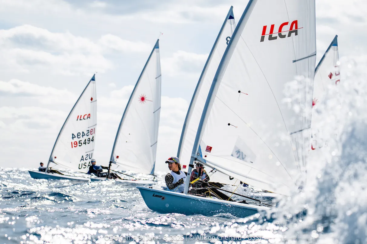 Several small sailboats with white sails, marked with ‘ILCA,’ racing on open water under a partly cloudy sky. The boats are angled slightly as they cut through choppy waves, with splashes of water in the foreground and sunlight reflecting off the sea surface