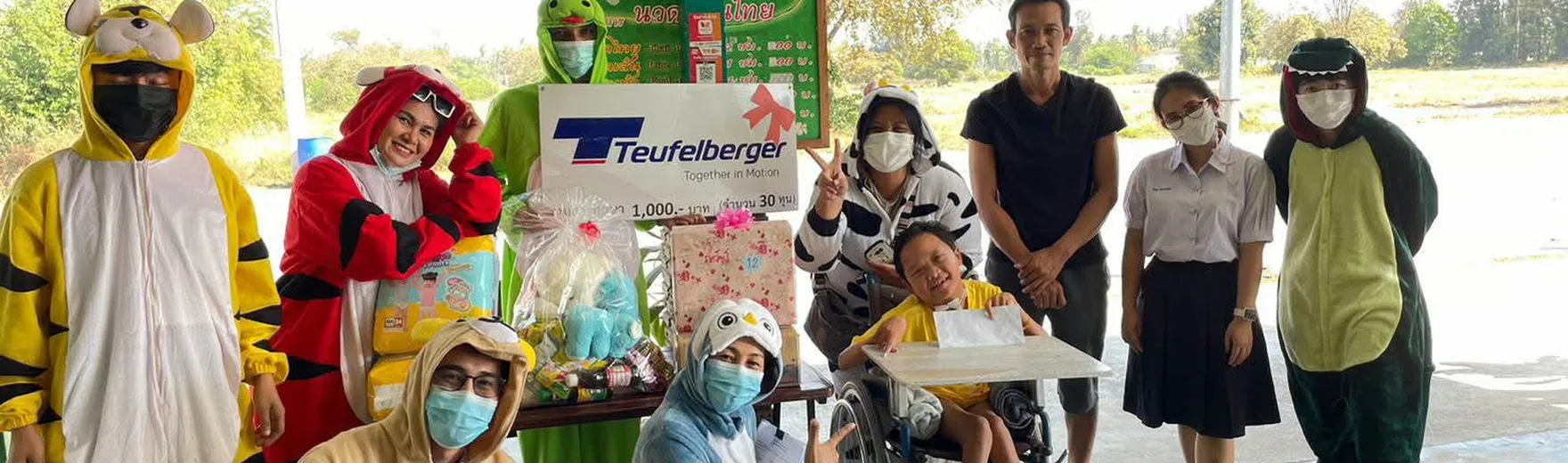 Group of people, some in animal costumes, pose with a child in a wheelchair holding a large check, under a covered outdoor area.