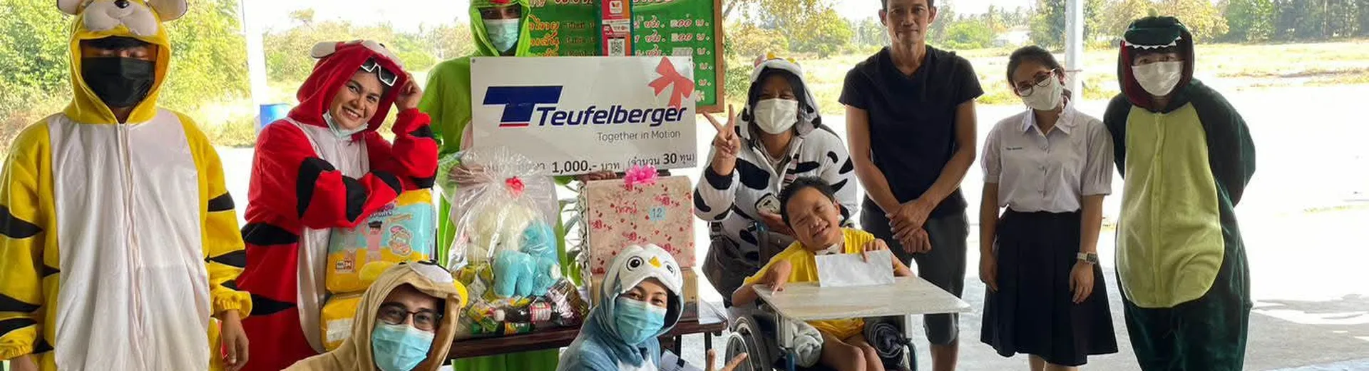 Group of people, some in animal costumes, pose with a child in a wheelchair holding a large check, under a covered outdoor area.