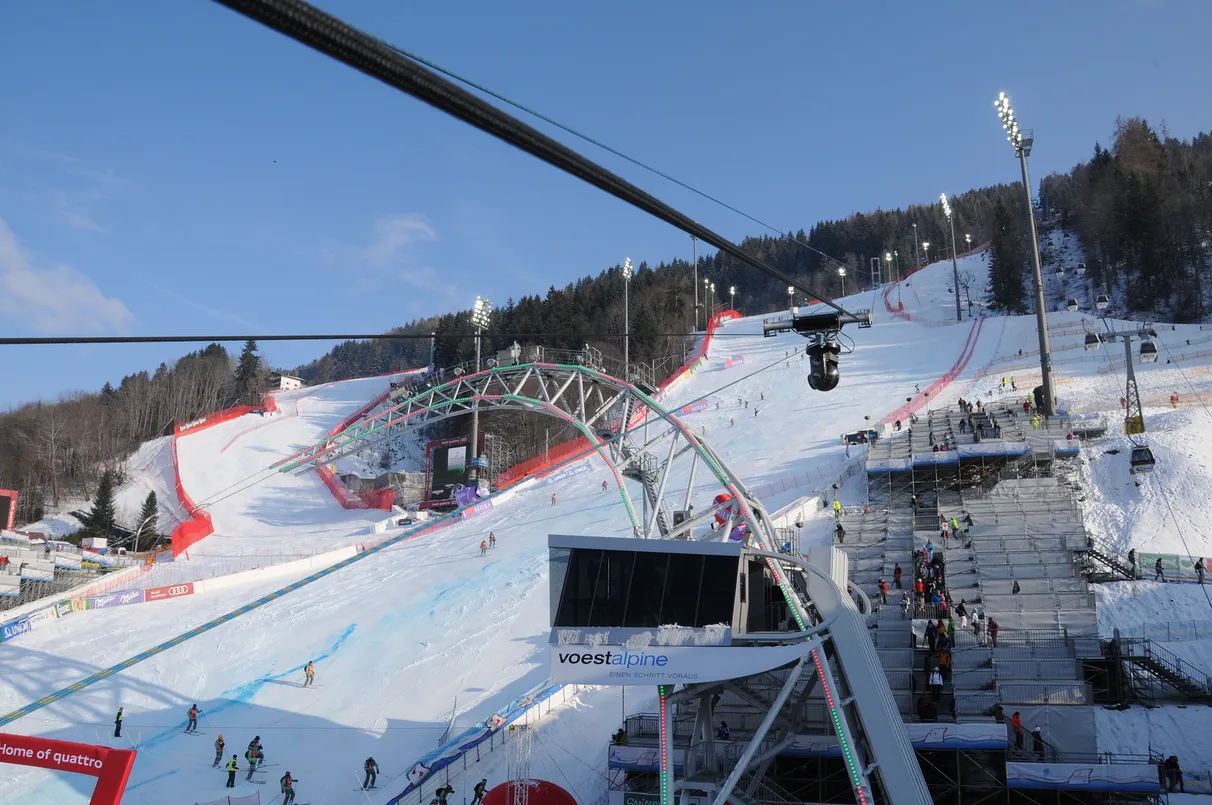 Ski slope with racers and spectators, surrounded by snowy mountains and trees, under a clear blue sky.
