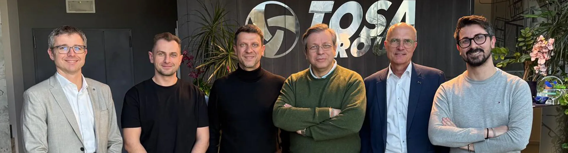 A group of six men standing in an office setting, smiling and posing for a photo in front of a sign with the words "TOSA GROUP."