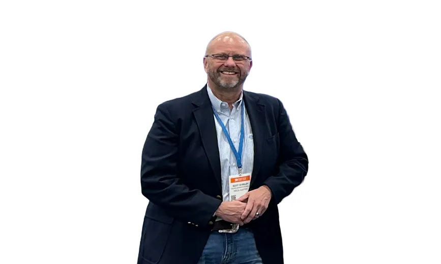 Man in a blazer with a conference badge, smiling in front of a plain white background.