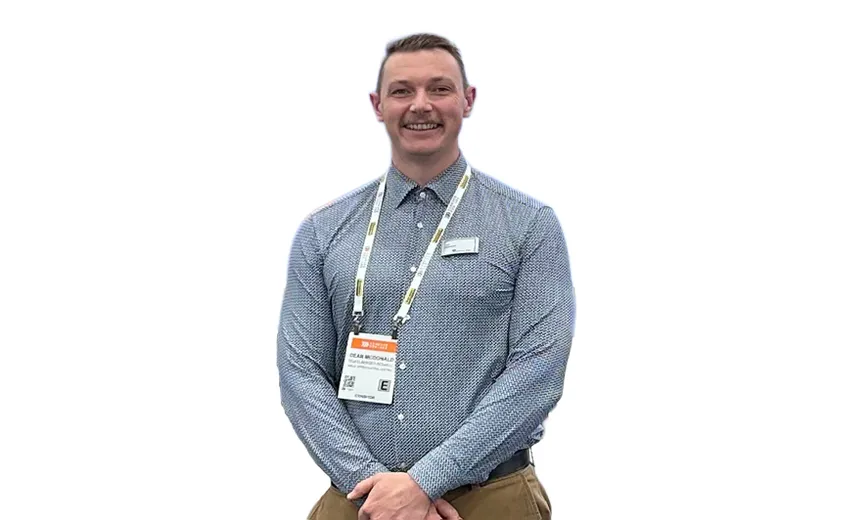 Man in a blue patterned shirt and lanyard smiles, standing against a white background.