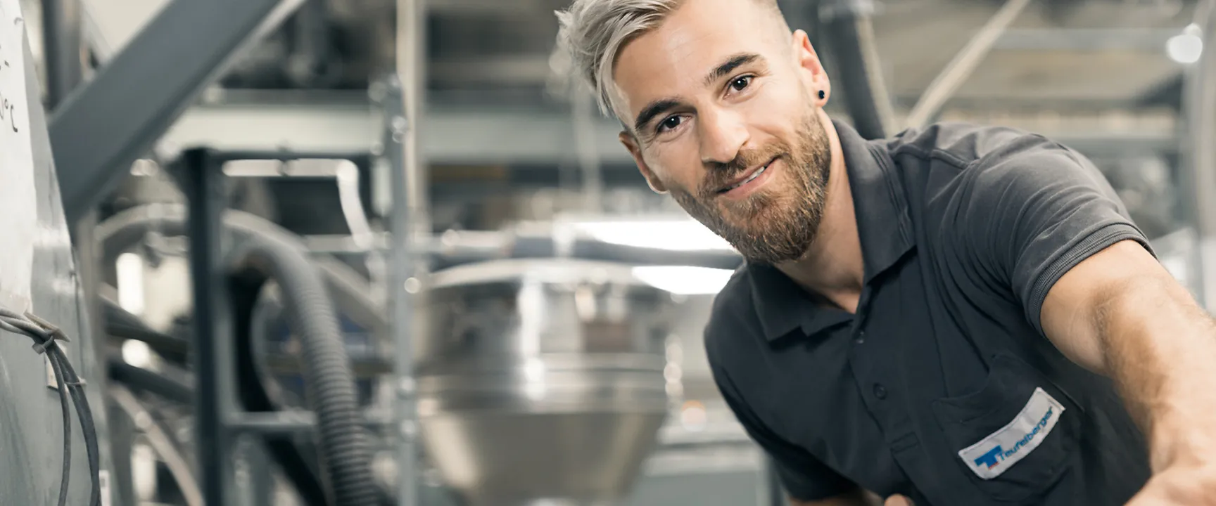 A person in a factory setting, wearing a gray shirt, smiling, with industrial machinery in the background.