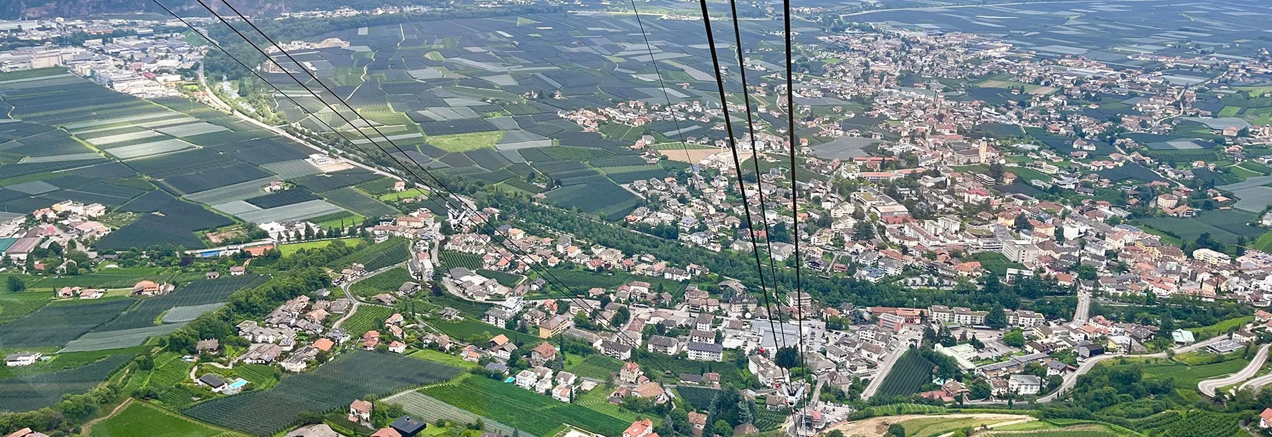 Aerial view of a lush valley with a patchwork of fields, a town, and distant mountains under a partly cloudy sky. Cable lines cross the scene.