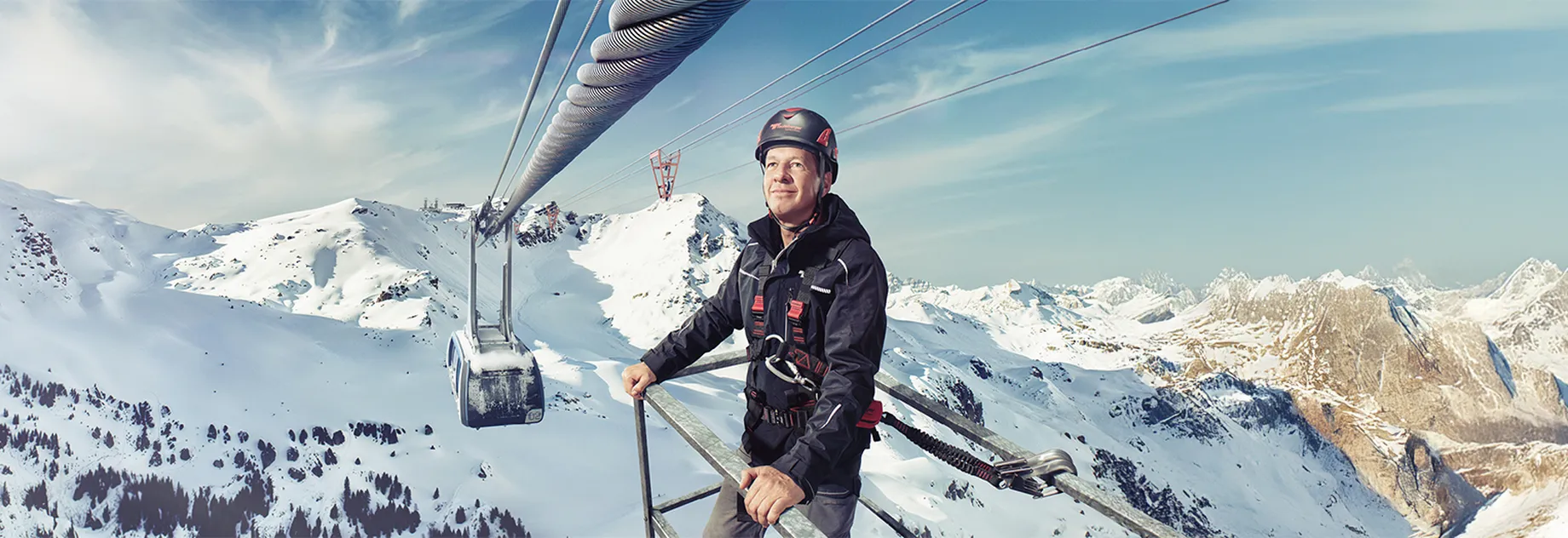 Technician in safety gear on alpine platform overseeing cable car system above snow-covered mountains under clear sky