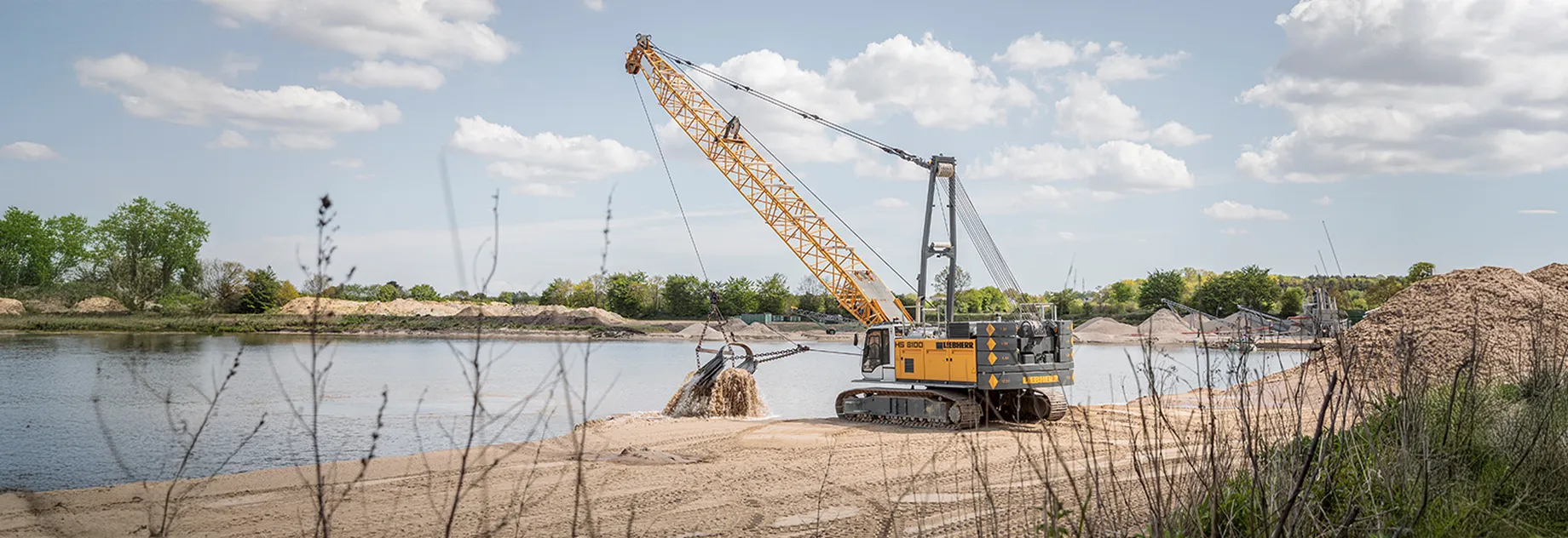 A Liebherr HS 8100 duty cycle crawler crane operating by a lakeshore, lifting sand from the water with a grab. Large sand piles and green trees in the background under a blue sky.
