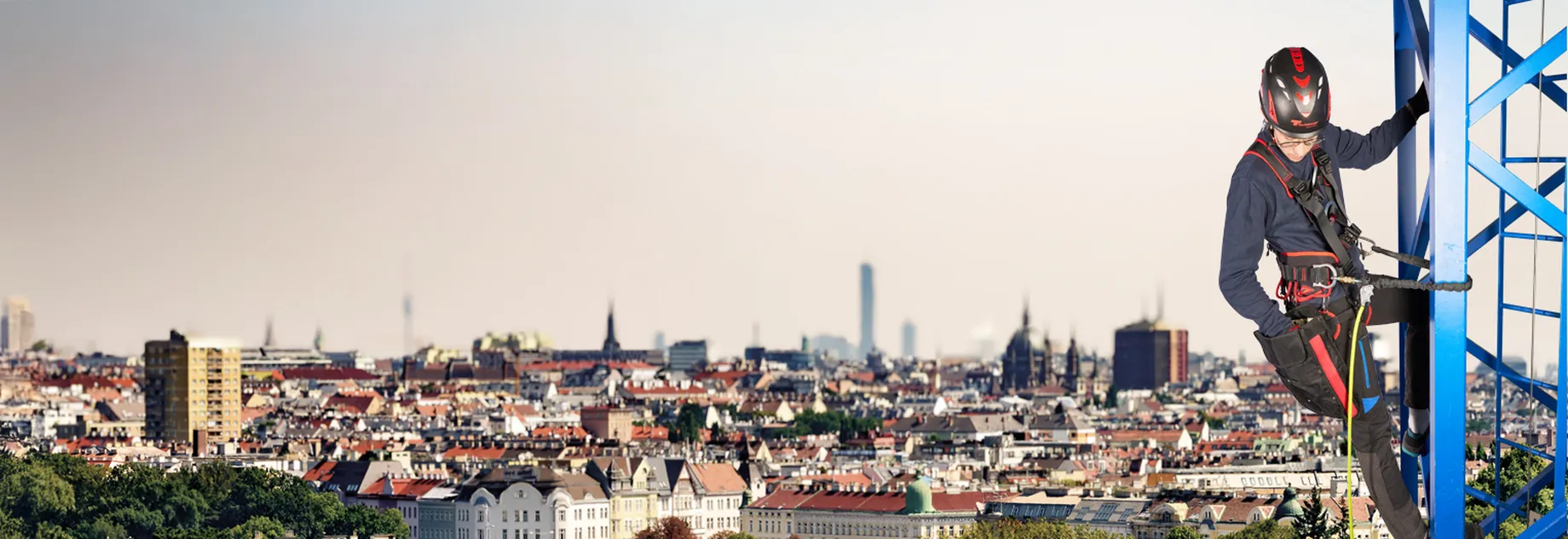 An industrial climber wearing a helmet and climbing equipment ascends a blue metal structure with a panoramic view of a city featuring high-rise and residential buildings under a clear sky.