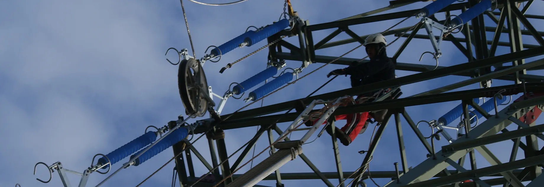 A worker in safety gear climbs a metal tower structure against a blue sky.