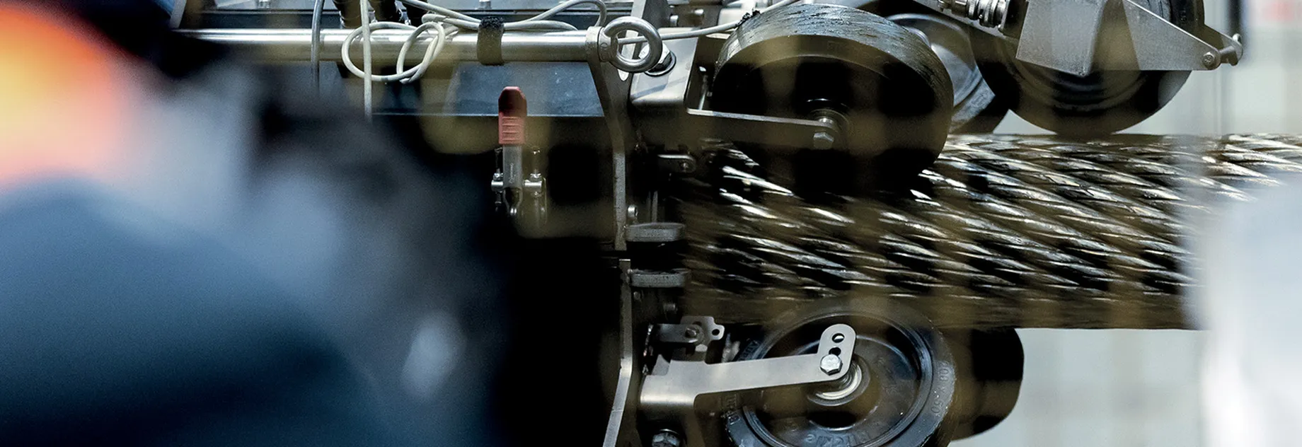 Close-up of industrial machinery processing a thick, steel wire rope, with gears and rollers in motion. Blurred foreground elements.