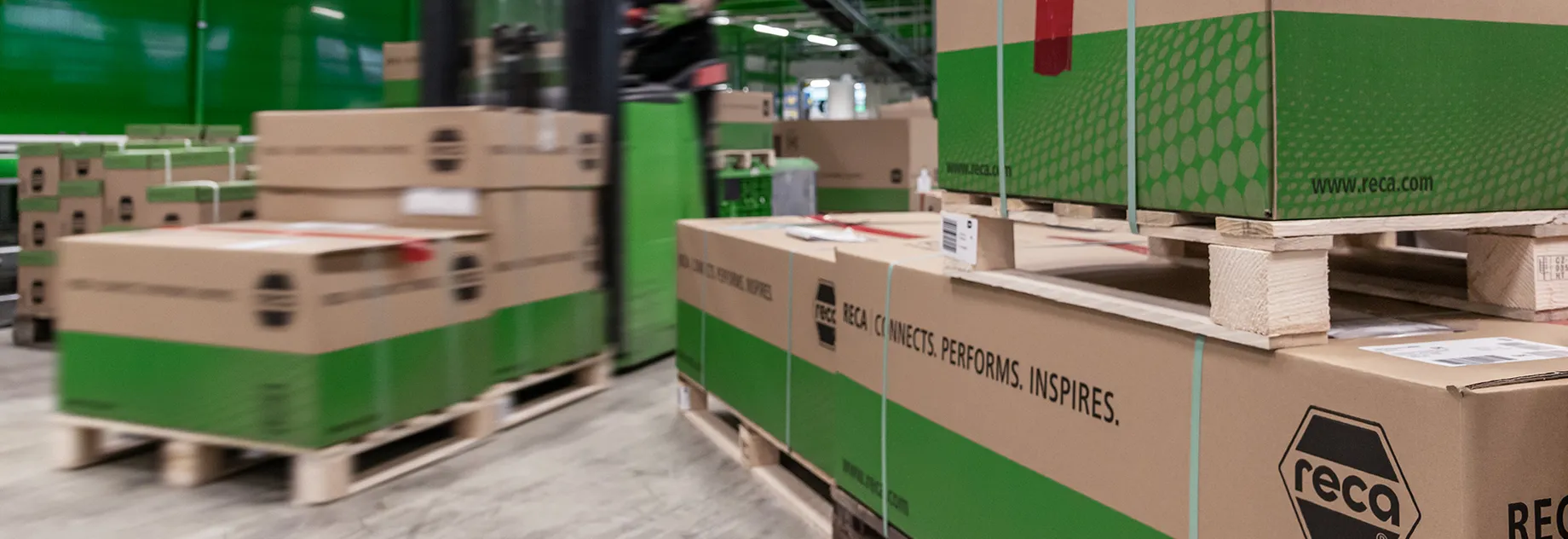 Warehouse area with several cardboard boxes stacked on wooden pallets; boxes feature the branding “reca” along with the slogan “RECA CONNECTS. PERFORMS. INSPIRES.” and the website www.reca.com.