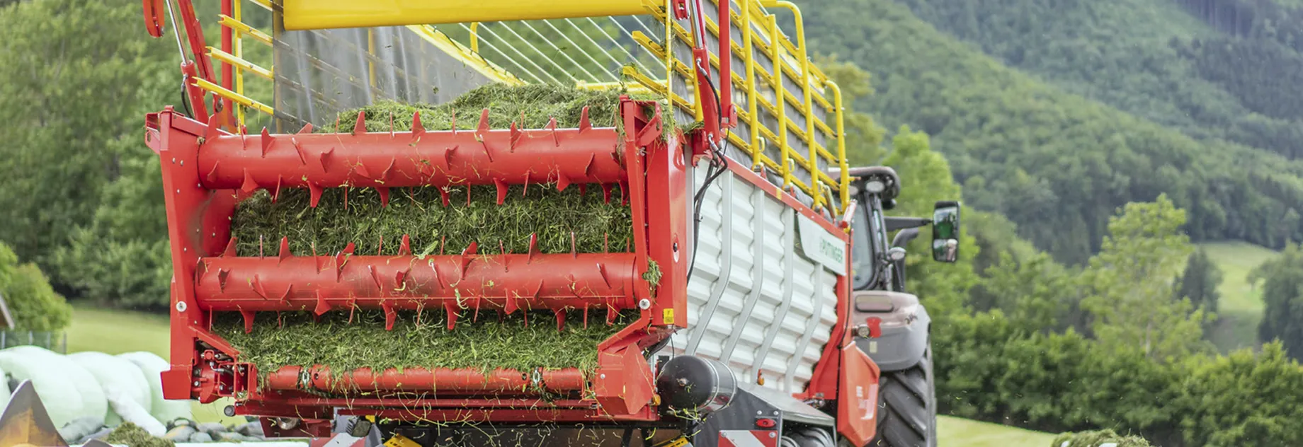 A red agricultural machine collects grass in a rural field, with green hills and trees in the background.