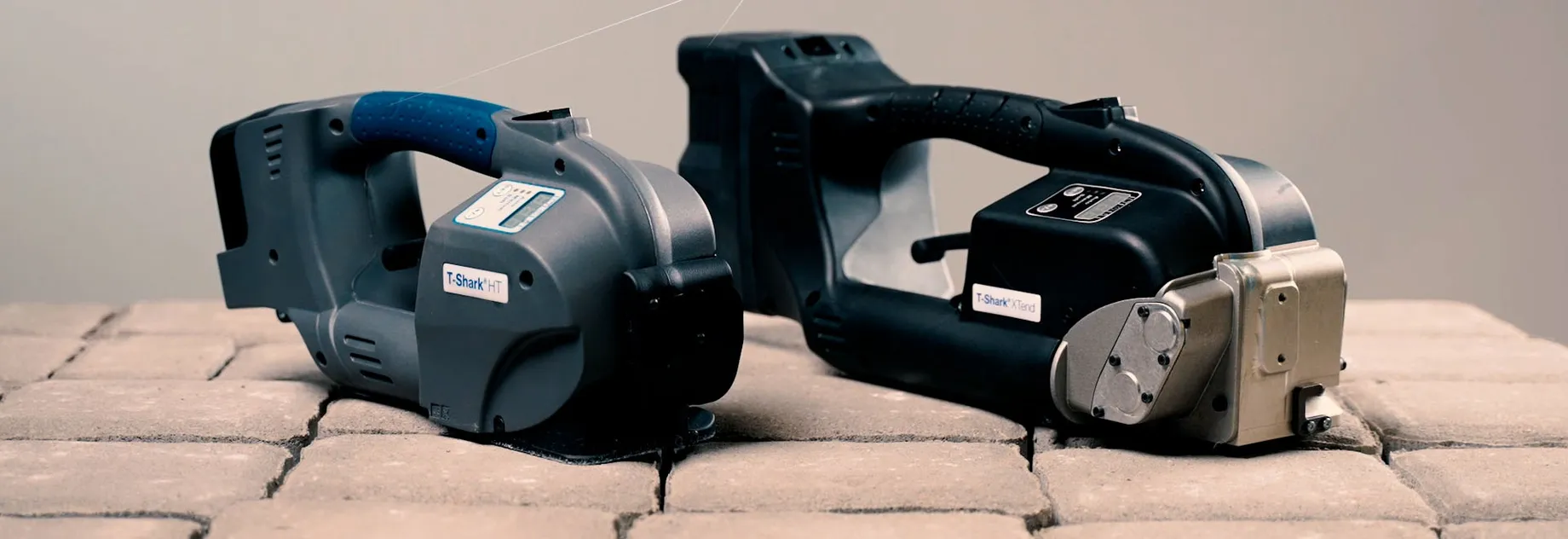 Two T-Shark tools placed on a surface made of paving stones. The left device is gray with a blue handle, the right one is black with a metallic front.