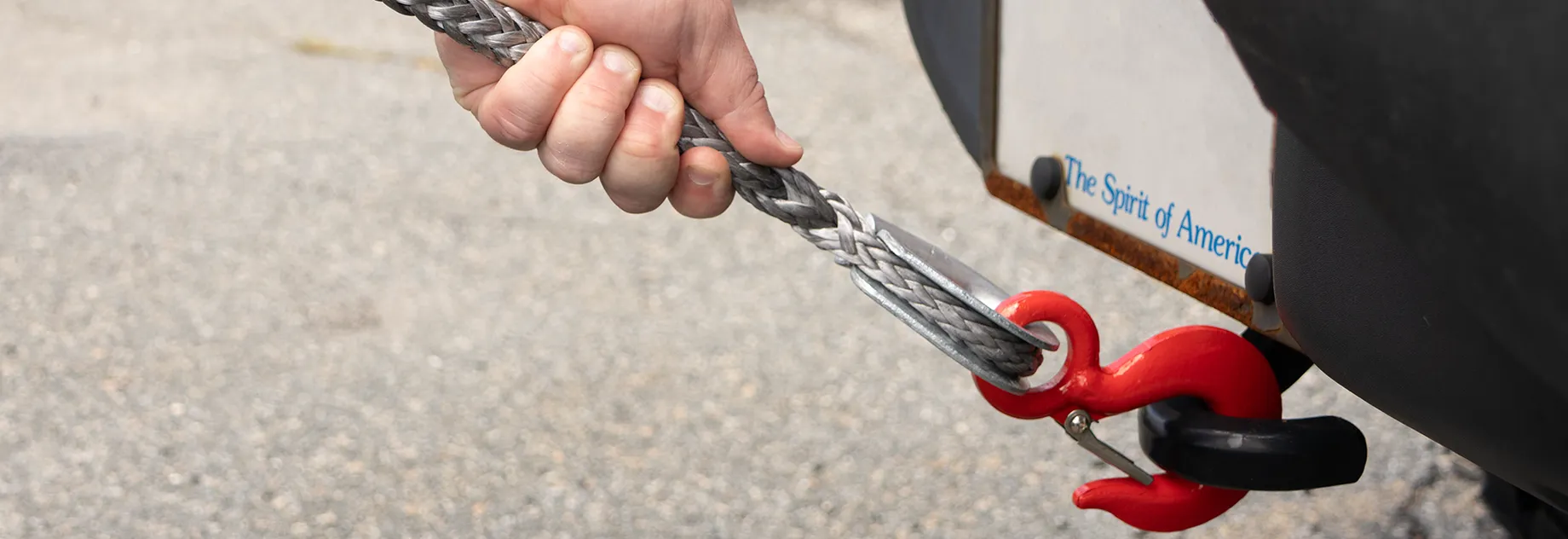 A hand holding a metal cable attached to a red hook, with a vehicle bumper in the background.