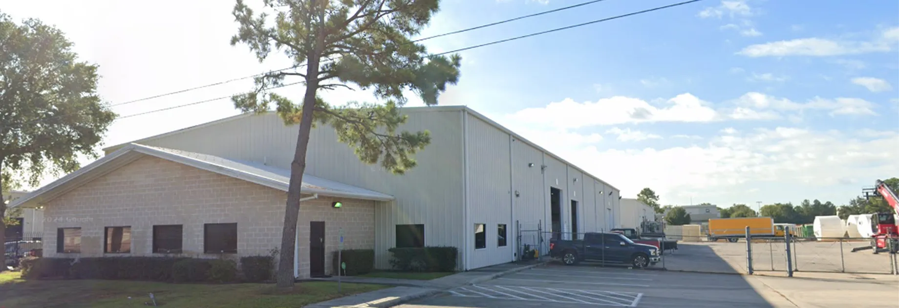 Warehouse building with a light-colored facade, surrounded by trees and parked vehicles, under a blue sky with clouds.