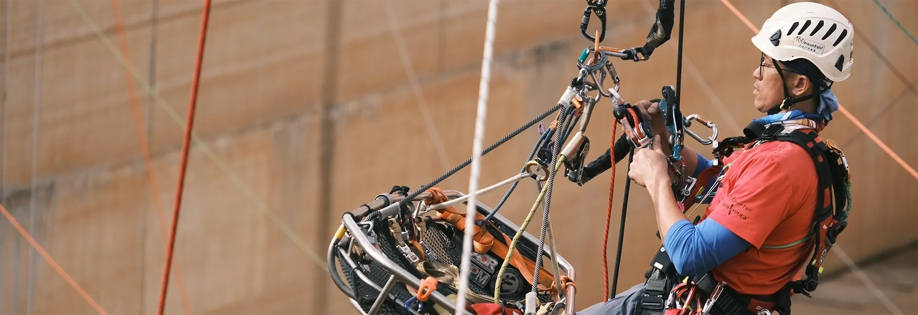A person in climbing gear and a helmet ascends using ropes and pulleys against a large wall.