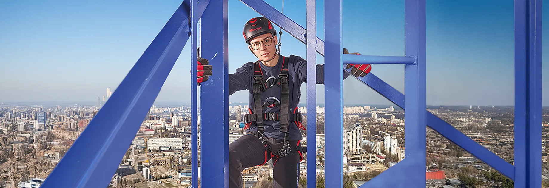 Industrial climber wearing safety gear working on a tall steel structure with a cityscape in the background.