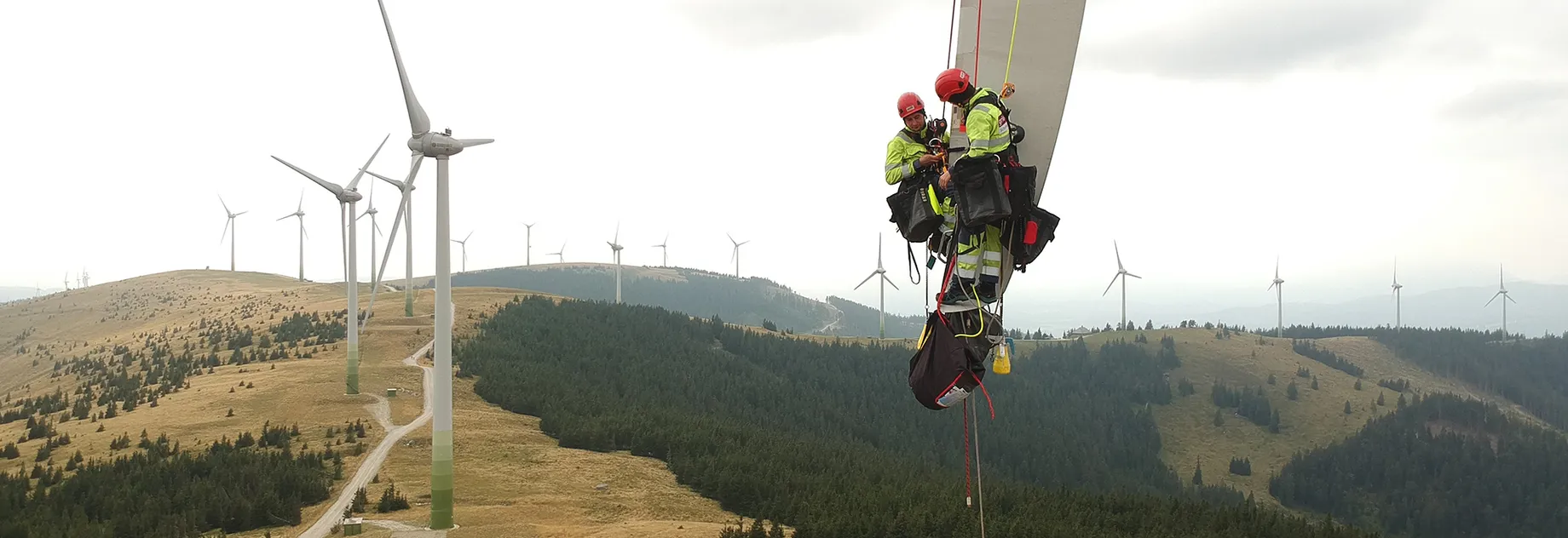 Fiber rope Two technicians in safety gear perform maintenance on a wind turbine blade, with multiple wind turbines visible on a hilly landscape.