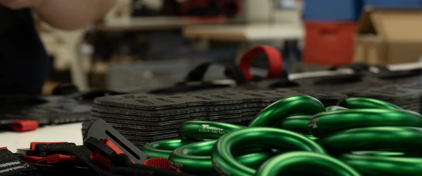 Close-up of a workshop workbench with green metallic carabiners in the foreground. Behind them are black plates with red straps, and blurred figures and tools in the background suggest an active workspace.