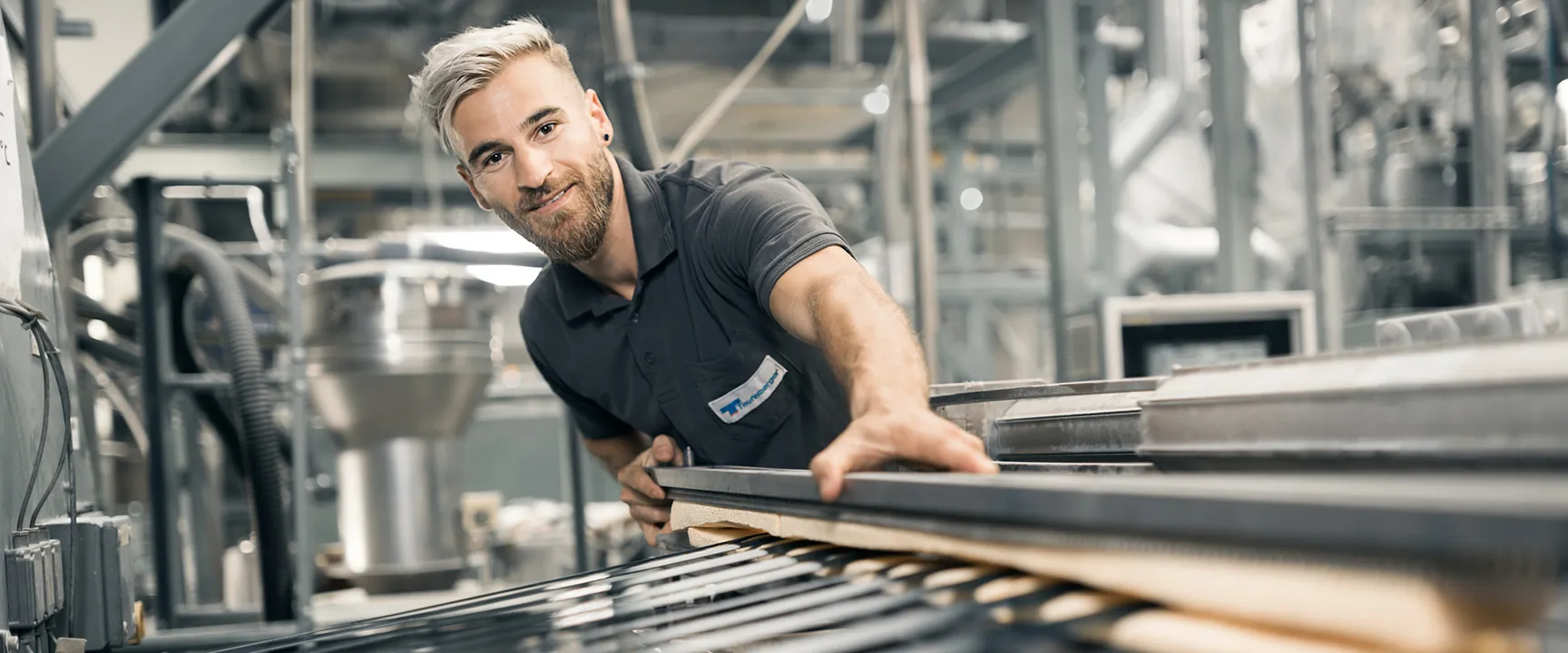 Person wearing a dark polo shirt with a name tag working in an industrial setting; face blurred for privacy, with machinery, pipes, and metal structures in the background.