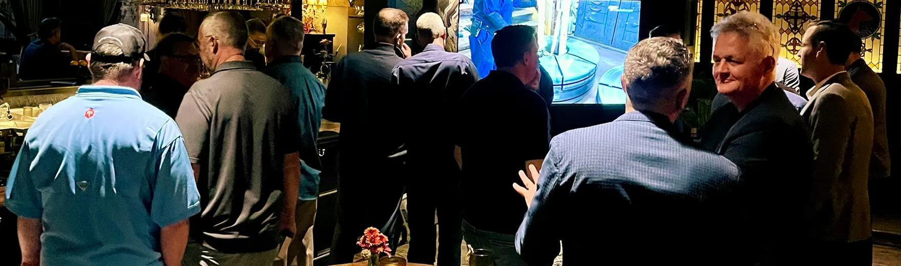 Two people standing outdoors next to a white signboard with a blue panel. In the background, there is a sidewalk, trees, a fence, and a building. The scene is captured in daylight with shadows cast on a brick wall.