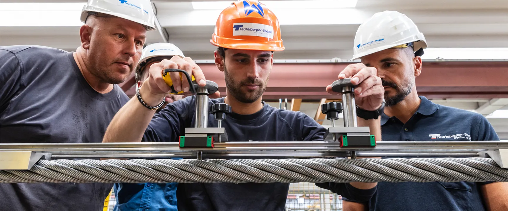 Workers in hard hats and uniforms inspect a large metal cable using measuring tools in an industrial setting.