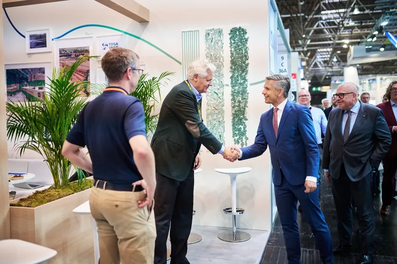 Two business professionals shaking hands at a trade show, surrounded by attendees and green decor.