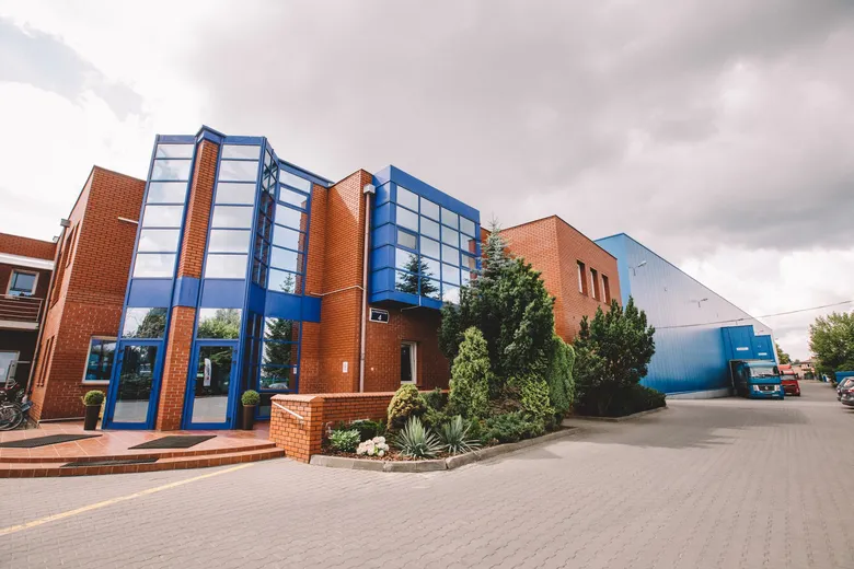 Modern industrial building with red brick and blue glass facade, surrounded by greenery, under a cloudy sky.