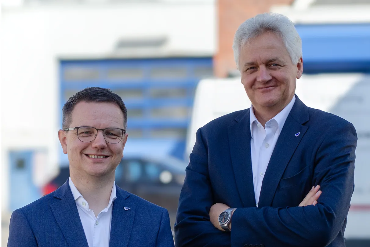 Two men in business attire stand near a wall-mounted screen displaying "1400." They are smiling and looking up at the camera.
