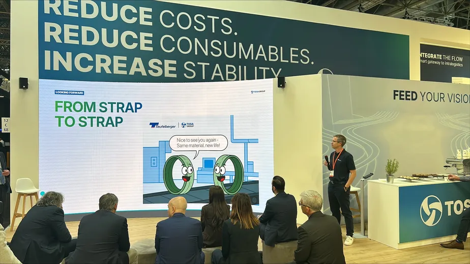 Presentation booth showcasing strapping solutions with attendees seated, highlighting themes of cost reduction and stability improvement.