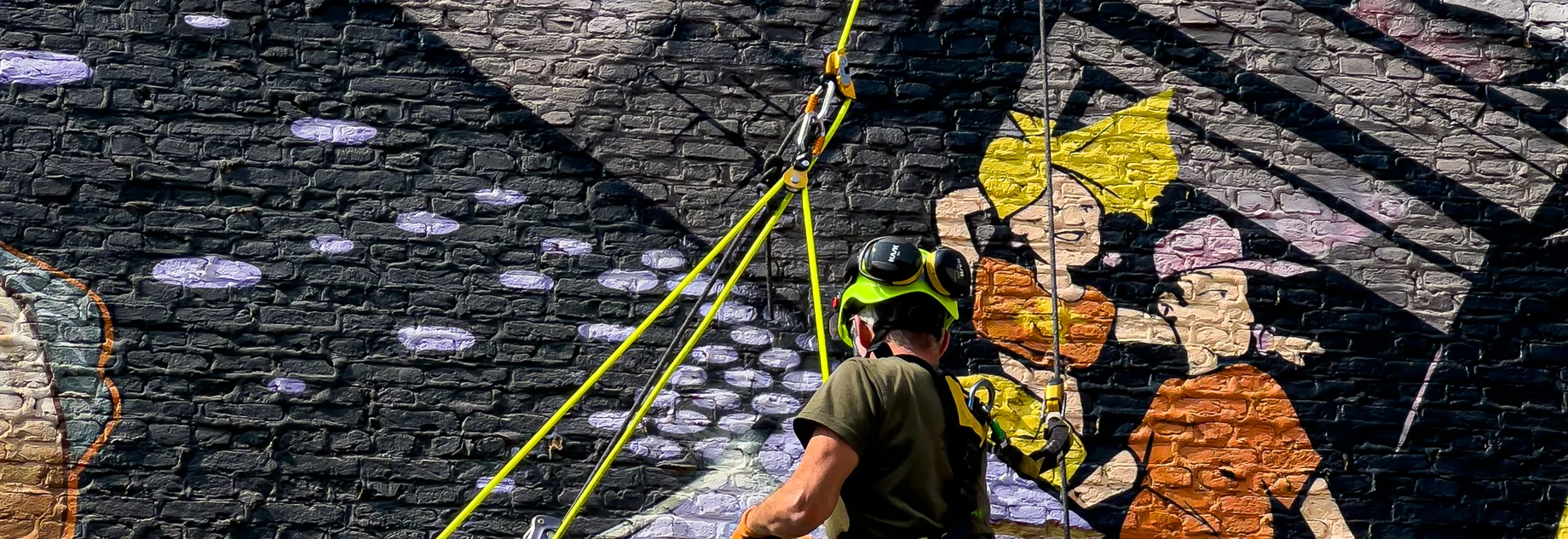 Rope Access worker working on a grafiti brick wall, while hanging on a high visible yellow ultrastatic rope. He is wearing a bright green helmet with ear protections folded back