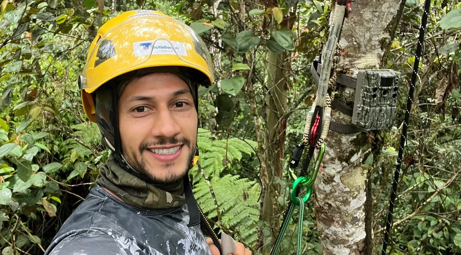 Science Ambassador Mateo is wearing a bright yellow helmet with a Teufelberger sticker on it while climbing a tree. On the stem of the tree he just installed a wildlife camera
