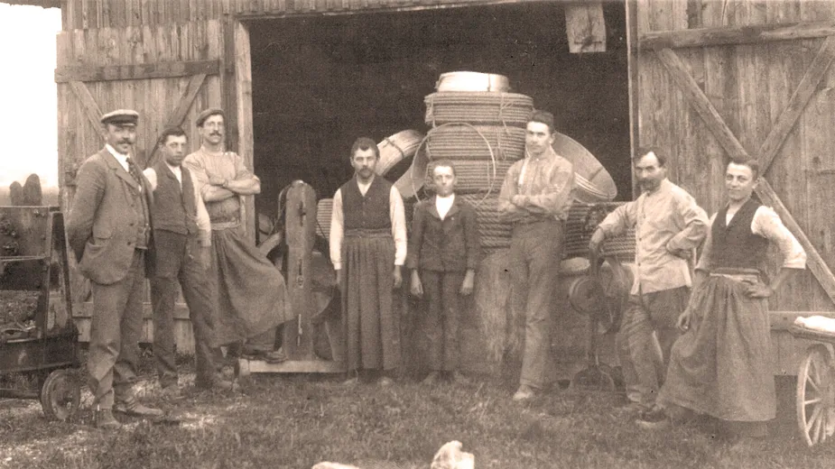 A vintage photo of eight people standing in front of a barn, with large barrels inside. They are dressed in early 20th-century attire.