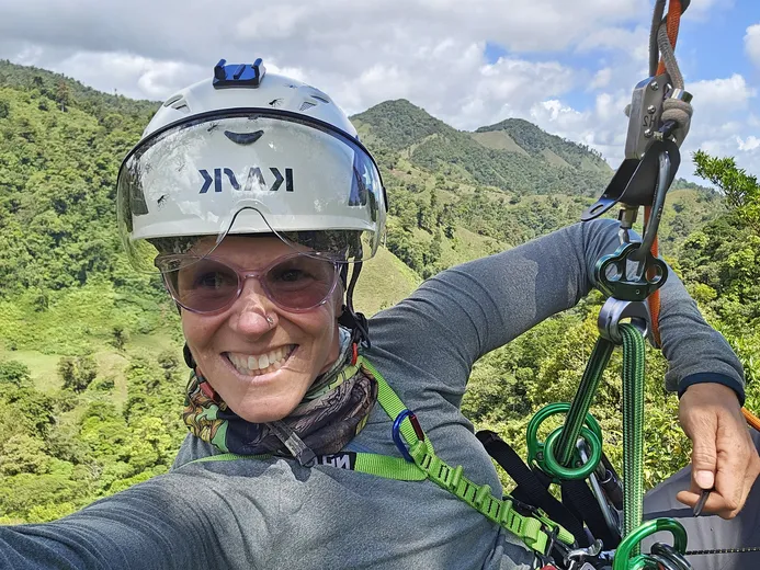 Selfie of Sybil hanging in a tree with a beautiful landscape in the back. She is wearing a white Kask helmet and sunglases.