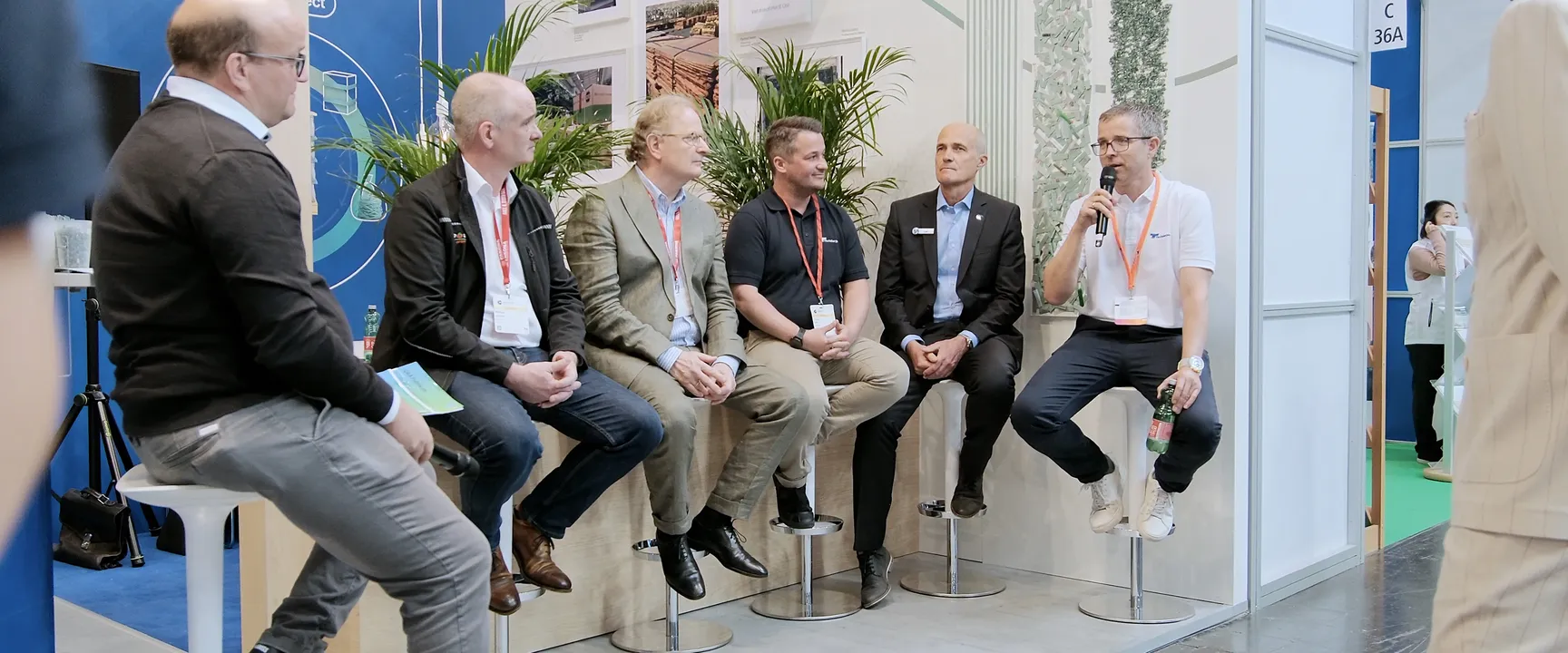Panel discussion with seven business professionals seated on stools, engaging in a conversation at a trade show booth.