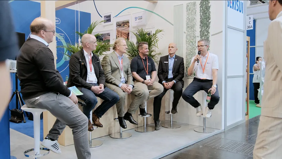 Panel discussion with seven business professionals seated on stools, engaging in a conversation at a trade show booth.