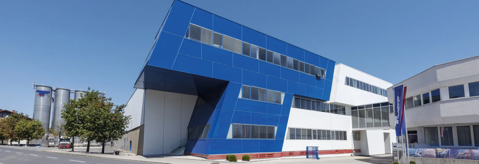 Modern blue and white industrial building with large windows, adjacent to a street and trees, under a clear blue sky.
