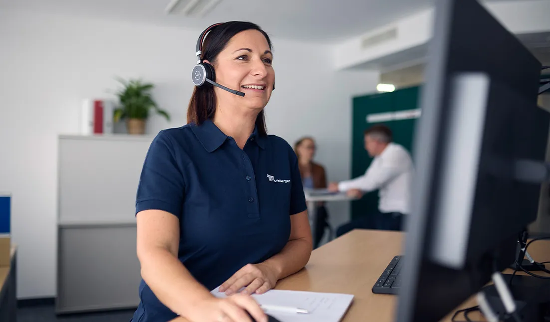 A person wearing a white shirt and headphones sitting at a desk with computer monitors in a modern office environment. The background includes windows with blinds and part of another workstation.