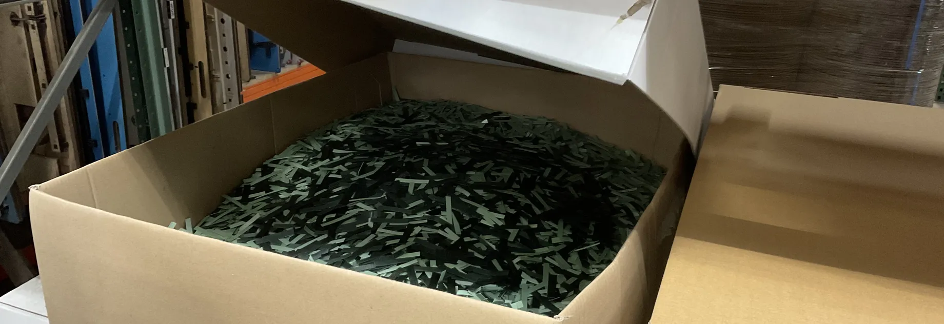Open cardboard box filled with green plastic strapping material on a warehouse shelf.