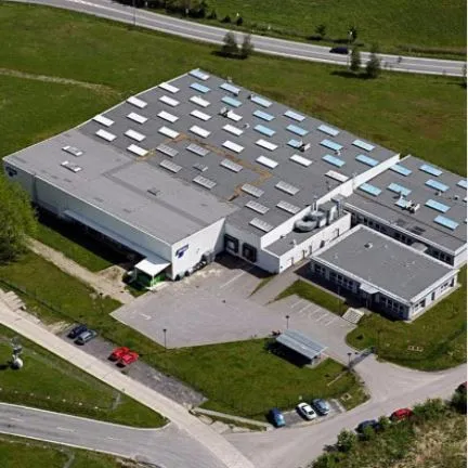Aerial view of a large industrial building with a flat roof featuring skylights and ventilation units. The facility is surrounded by green fields and located near a road, with several cars parked in the adjacent lot.
