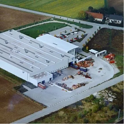 Aerial view of an industrial facility featuring a large white building with multiple sections and loading docks. The surrounding area includes parked vehicles, storage zones, and nearby fields, with a road providing access to the site.