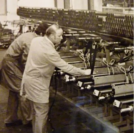 Two individuals in lab coats working at a long workbench in a factory setting, assembling or operating machinery with visible cables and components. The background shows additional industrial equipment.