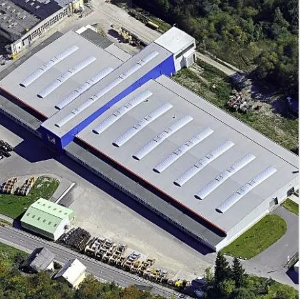 Aerial view of a large industrial building with a T-shaped layout and a grey roof featuring multiple skylights. The site includes paved areas, greenery, nearby structures, and visible vehicles and equipment.
