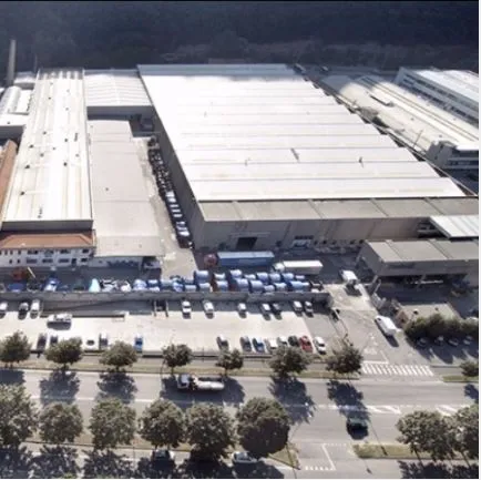 Aerial view of a large industrial complex with multiple warehouses, parked trucks, and cars, surrounded by roads and trees.