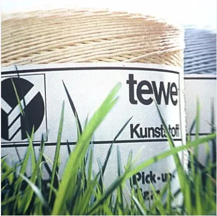 Close-up of a spool of synthetic twine labeled 'tewe Kunststoff Pick-up', partially obscured by grass blades in the foreground. The label features geometric shapes and indicates the twine is made of plastic.